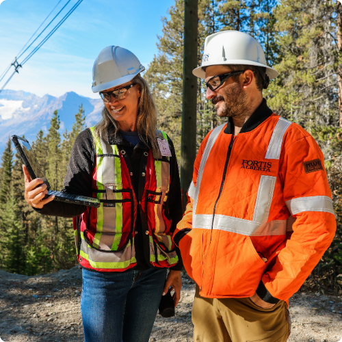 Female and male employee in safety gear reviewing a document outdoors, on a laptop