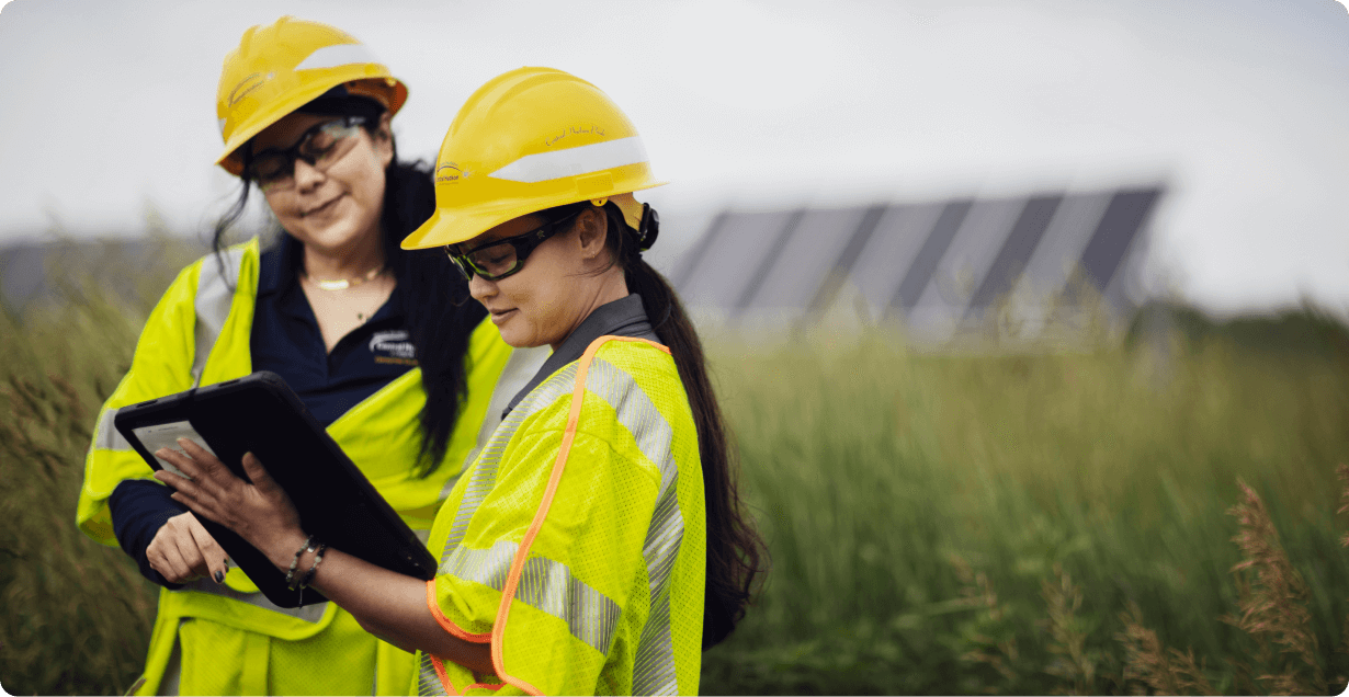 Two women in safety gear reviewing a tablet with solar panels in the background